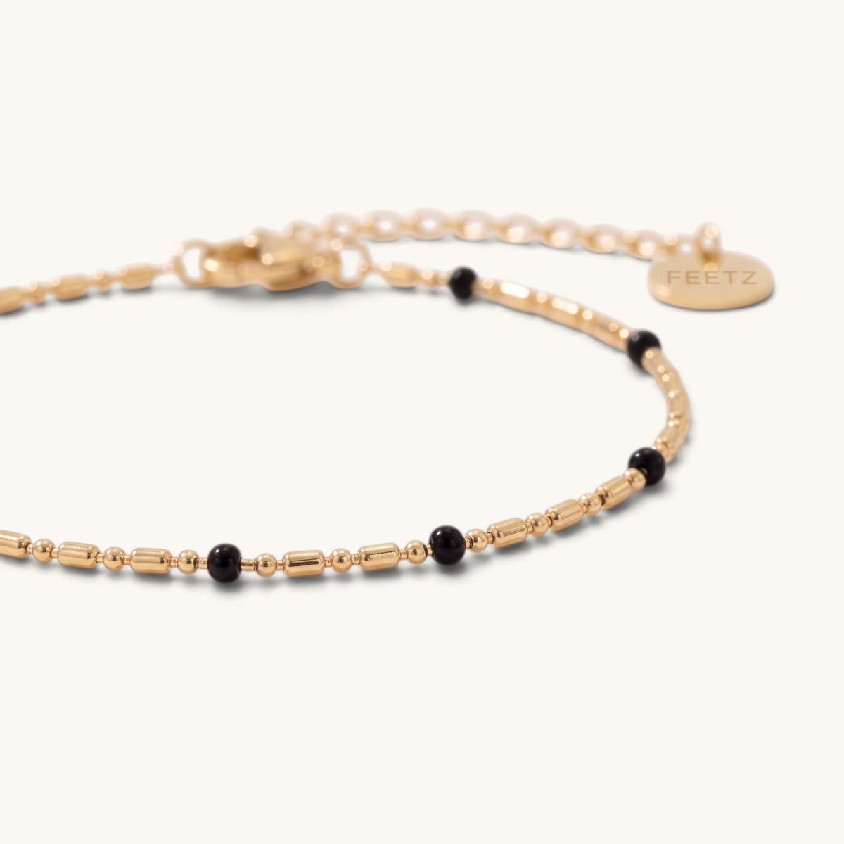 Feetz & Co. Onice gold stainless steel anklet with delicate chain and small black beads, featuring a logo charm.
